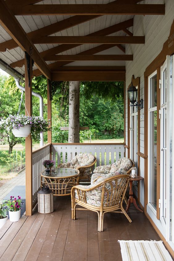 a small and pretty Scandinavian porch with rattan seating furniture with floral upholstery, a round rattan table and some blooms is summer-ready