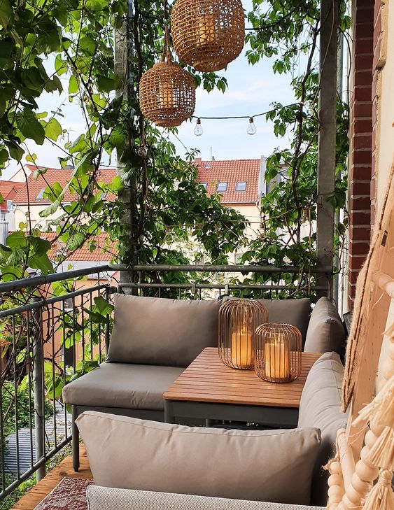 a small and welcoming balcony with upholstered furniture, a wooden table, greenery, woven pendant lamps and candle lanterns
