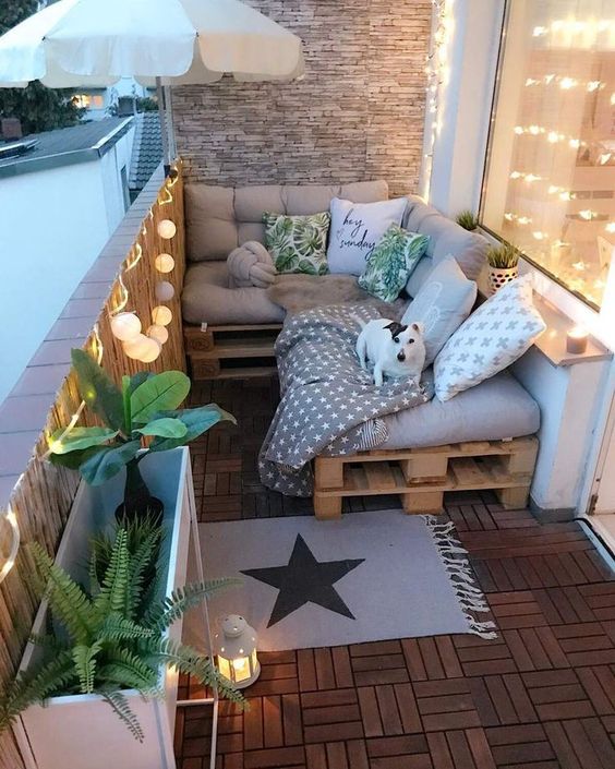 a small boho balcony with a pallet daybed, pillows, an umbrella, potted plants, string lights and candle lanterns