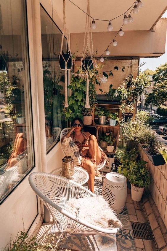 a small boho balcony with lots of potted plants, woven chairs and a table, string lighs and a boho rug