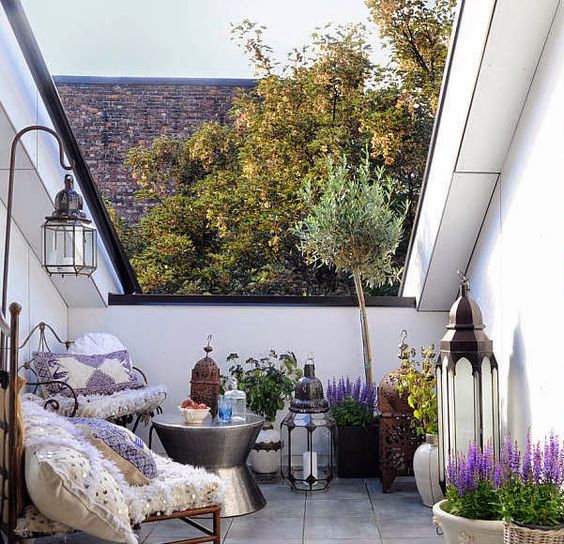 a small boho terrace with various candle lanterns, potted greenery and blooms and forged chairs with boho pillows