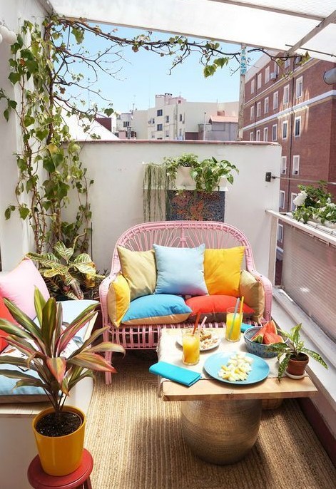 a small bright balcony with a pink loveseat, a small table, a white bench with storage, potted greenery and bright pillows