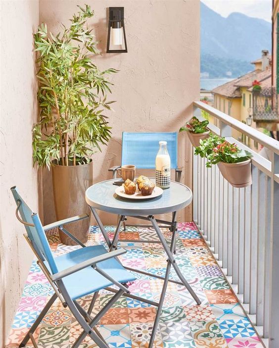 a small colorful balcony with bright tiles on the floor, a folding table and blue chairs, potted blooms and plants