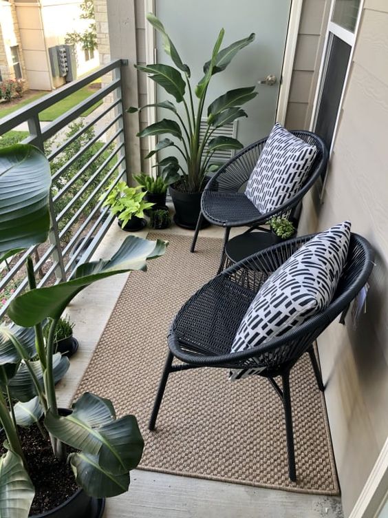 a small modern balcony with a jute rug, black chairs and pillows and some potted tropical plants
