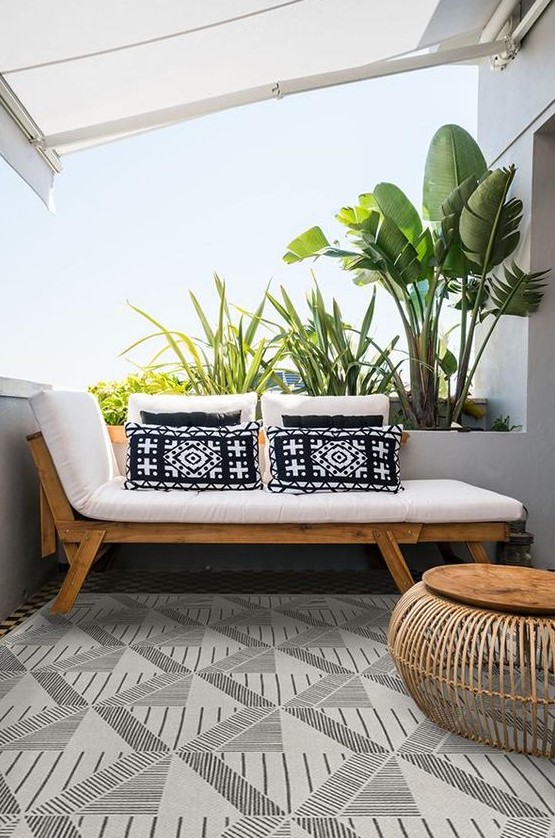 a small modern balcony with a tropical feel, with a lounger and printed pillows, a wooden coffee table and potted plants