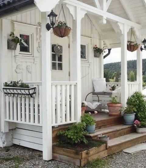 a small porch with little wooden benches with neutral pillows, potted plants and blooms is a welcoming and lovely space