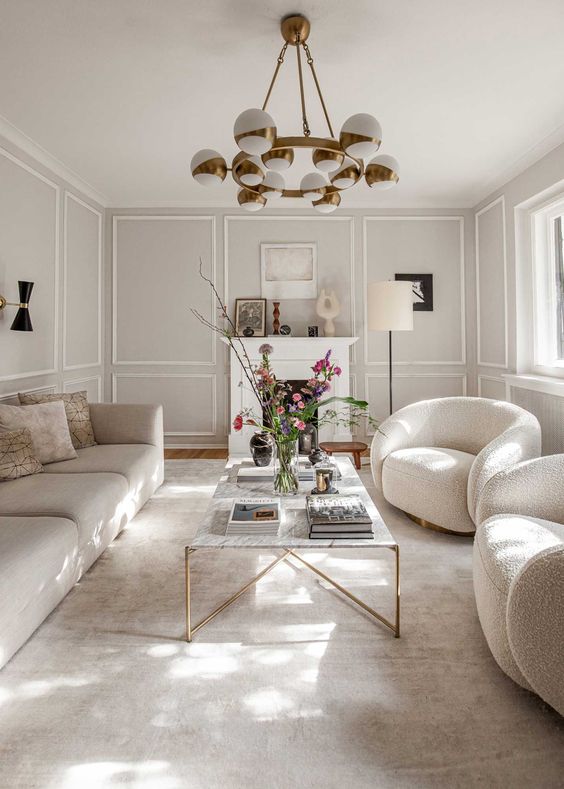 a sophisticated neutral living room with wainscoting, chic neutral furniture, a refined coffee table and a faux fireplace for more coziness