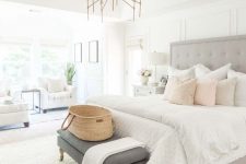 a stylish neutral bedroom with a grey upholstered bed, a brass chandelier, a grey bench, white chairs and pillows