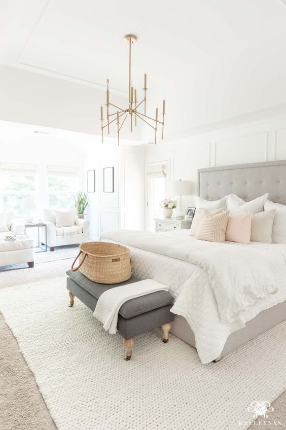 a stylish neutral bedroom with a grey upholstered bed, a brass chandelier, a grey bench, white chairs and pillows