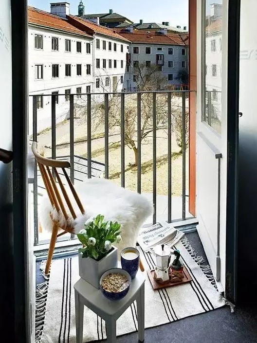 a tiny Nordic balcony with a wooden chair and a small white stool as a table, with blooms, a striped rug and a faux fur cover on the chair