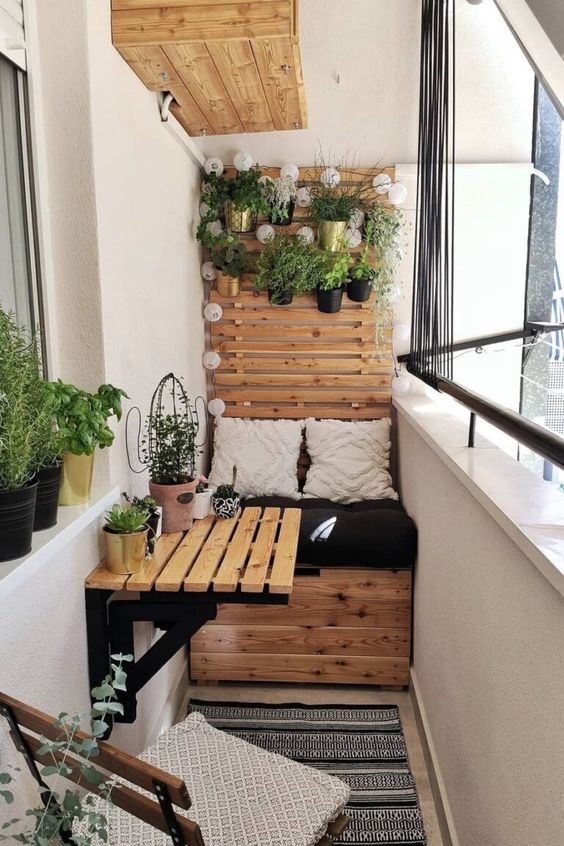 a tiny balcony with a built-in storage bench, a folding table and a chair, potted greenery and printed boho textiles