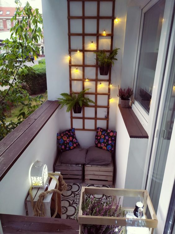 a tiny balcony with a pallet seat, a trellis with lights and potted greenery, a glass coffee table and a candle lantern
