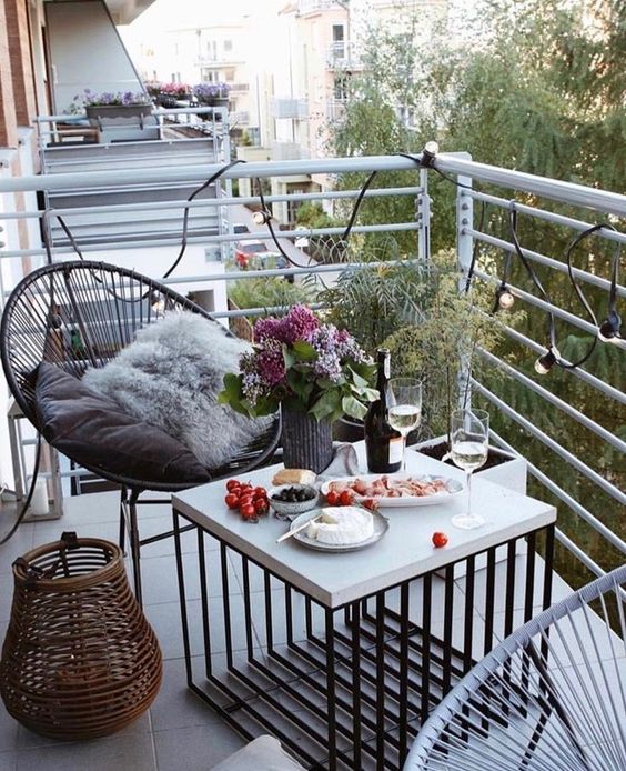 a tiny balcony with wicker chairs and a concrete coffee table, potted plants and blooms and string lights