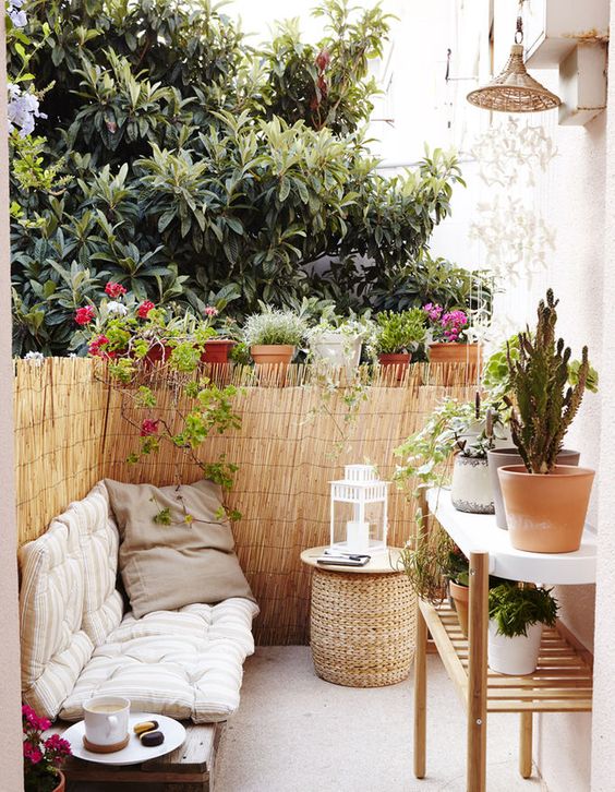 a tiny boho terrace with an upholstered bench, a wicker ottoman, a large plant stand and lots of potted plants around