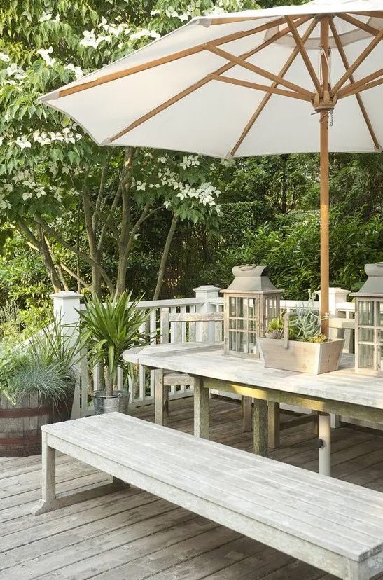 a very simple modern farmhouse terrace with a whitewashed dining set with benches, an umbrella and candle lanterns plus potted greenery