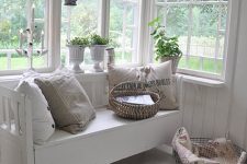 a white Nordic sunroom with a vintage bench, neutral pillows, a wire basket, potted greenery, a pendant lamp