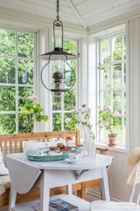 a white Scandi sunroom that is a dining room with stained and painted furniture, with lots of potted blooms and a vintage metal lamp