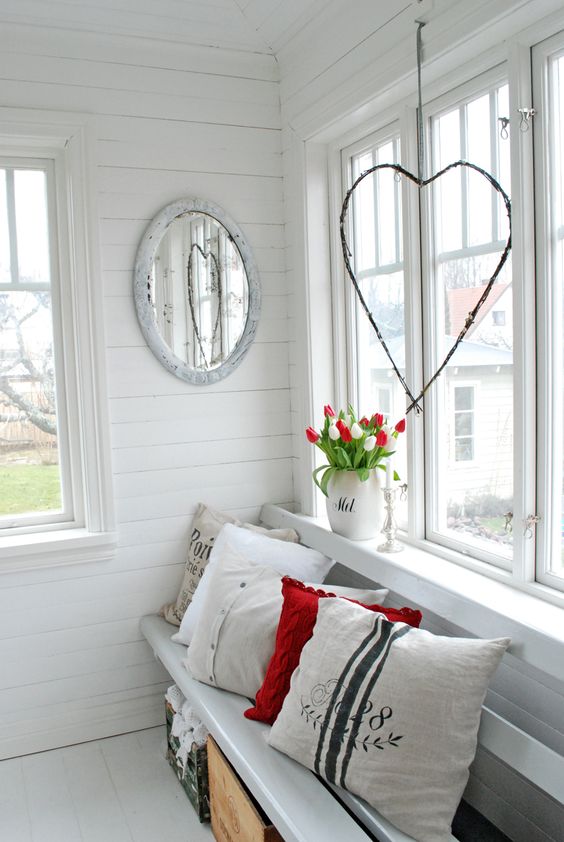 a white Scandi sunroom with planked walls, a built in bench with printed pillows, crates for storage, tulips in a jug and a heart