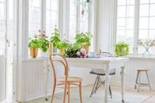 an airy white Scandinavian sunroom with a vintage dining table, vintage chairs, pendant lamps, potted plants and blooms