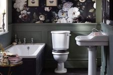 an exquisite Gothic bathroom with dark floral wallpaper and green panels, a tub with black panels, white appliances, a chic table and butterfly art