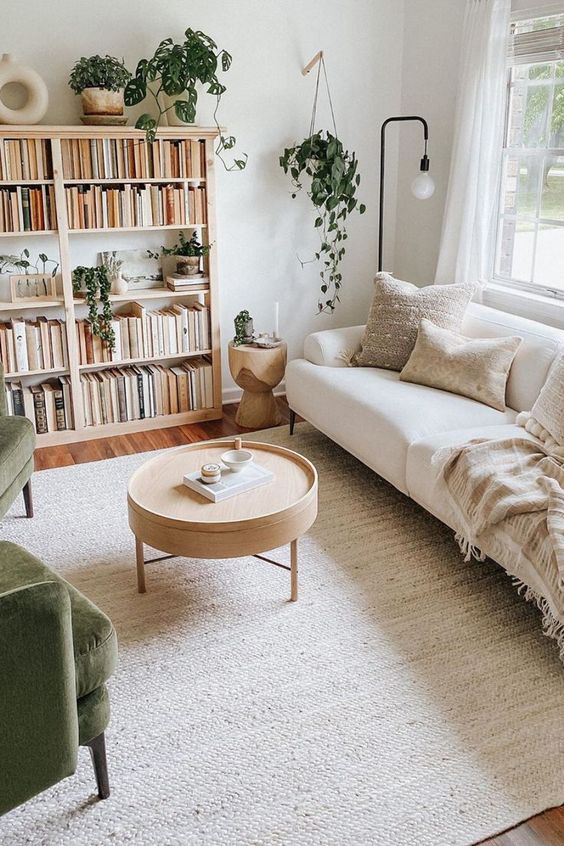 an inviting living room in neutrals, with a creamy sofa, a floor lamp, an open bookshelf, green chairs and a round coffee table
