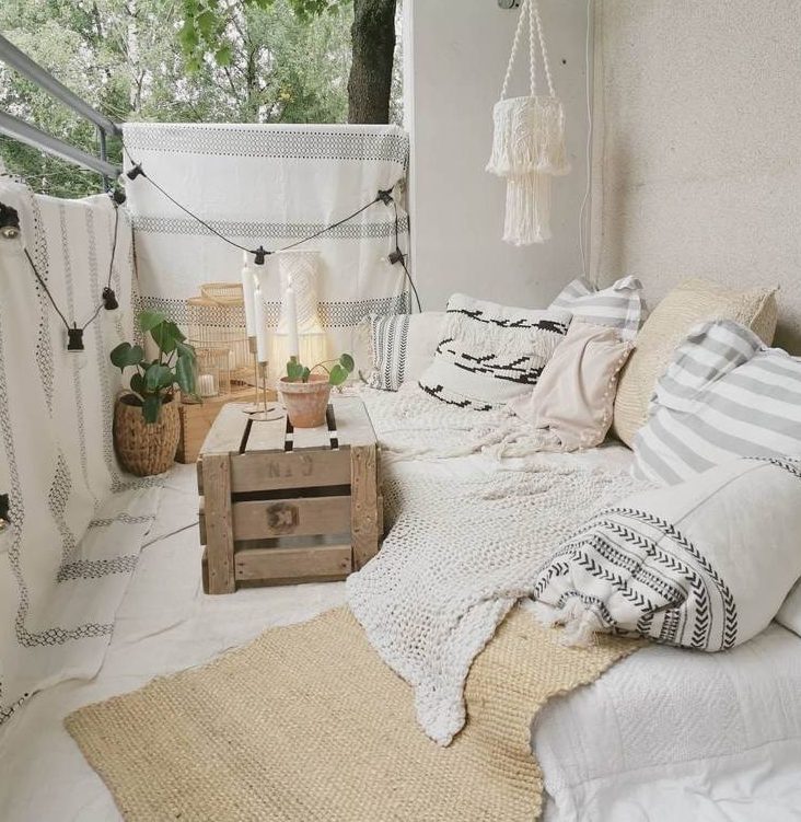 a beautiful creamy boho balcony with a low sofa with lots of pillows and blankets, a crate as a table, layered rugs and striped blankets hanging for privacy