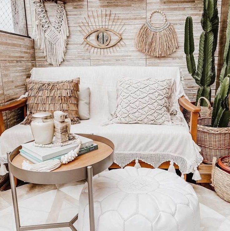 a beautiful neutral boho balcony with a sofa with boho pillows, a boho rug, a white pouf, potted plants, a side table and macrae and hangings of various kinds