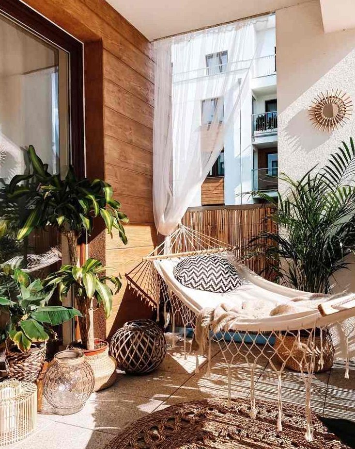 a boho balcony with a hammock and pillows, potted plants, candle lanterns, a jute rug and curtains, a sunburst mirror is a very cool and stylish idea to rock