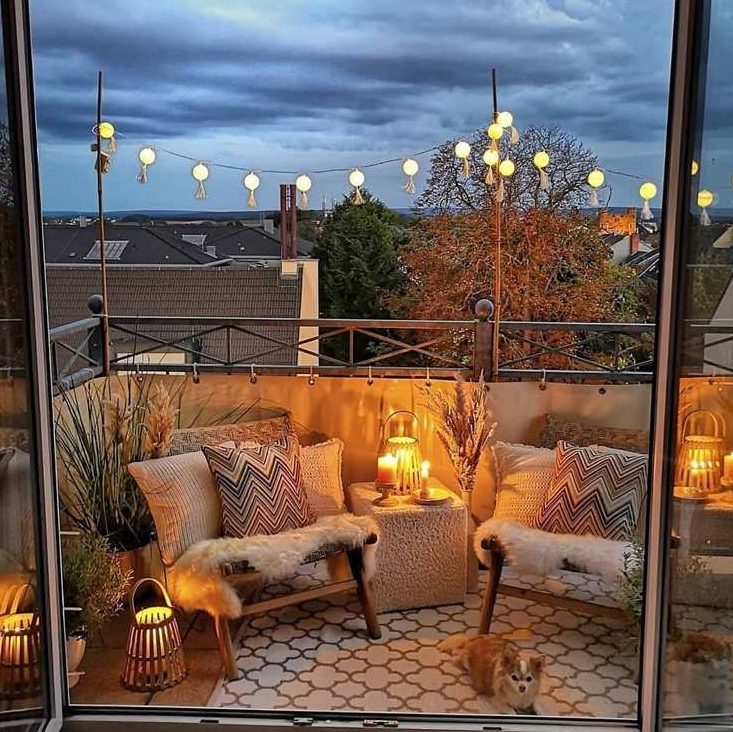 a boho balcony with a printed rug, neutral chairs with printed boho pillows, candle lanterns, potted plants and pampas grass in vases
