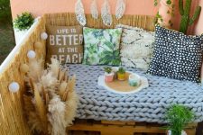 a boho balcony with black and white tiles, a pallet sofa with brigth pillows, a dream catcher, potted plants and a bit of pampas grass in a vase
