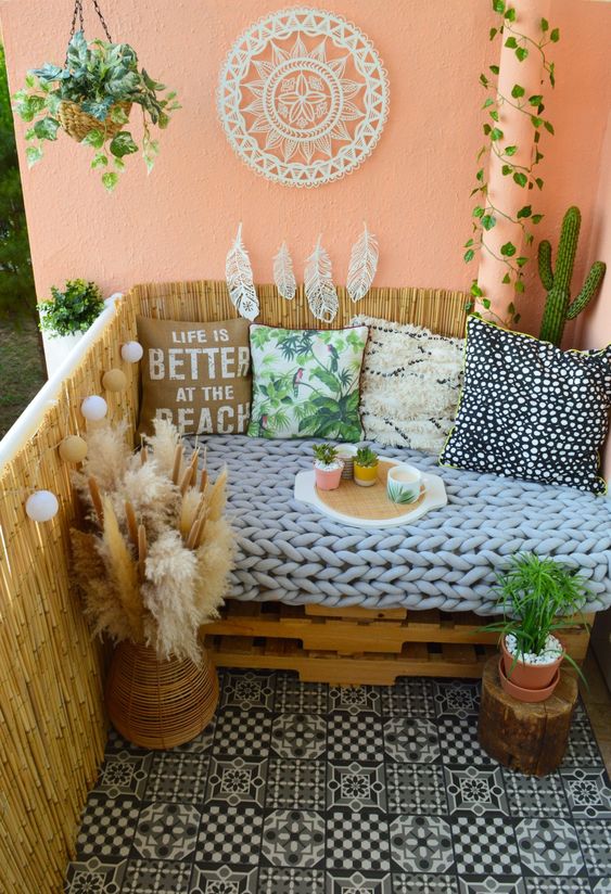 a boho balcony with black and white tiles, a pallet sofa with brigth pillows, a dream catcher, potted plants and a bit of pampas grass in a vase