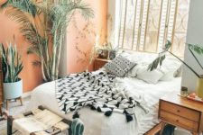 a bright boho bedroom with a rattan screen, wooden furniture, a peachy wall, potted plants and greenery and a wicker stool