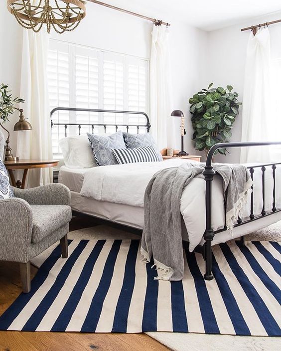 a bright vintage farmhouse bedroom with a metal bed, vintage furniture, grey and blue bedding, potted plants and a beaded chandelier