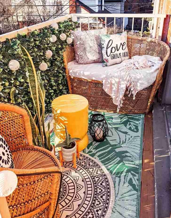 a colorful boho balcony with rattan seats, layered rugs, a yellow pouf, candle lanterns, bulbs and potted plants is a lovely and chic space to be