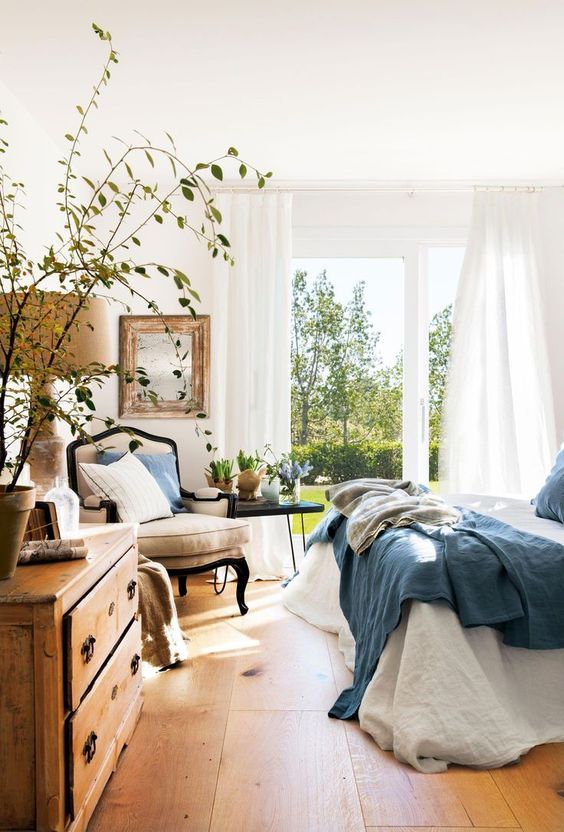 a country spring bedroom with vintage furniture, blue bedding, greenery and potted plants, a mirror in a wooden frame