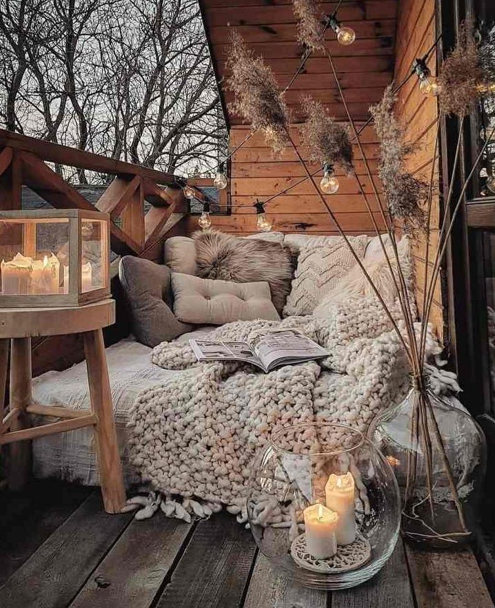 a cozy boho balcony with a daybed with lots of pillows and a blanket, lots of candles in various candle lanterns, some grass and bulbs over the bed