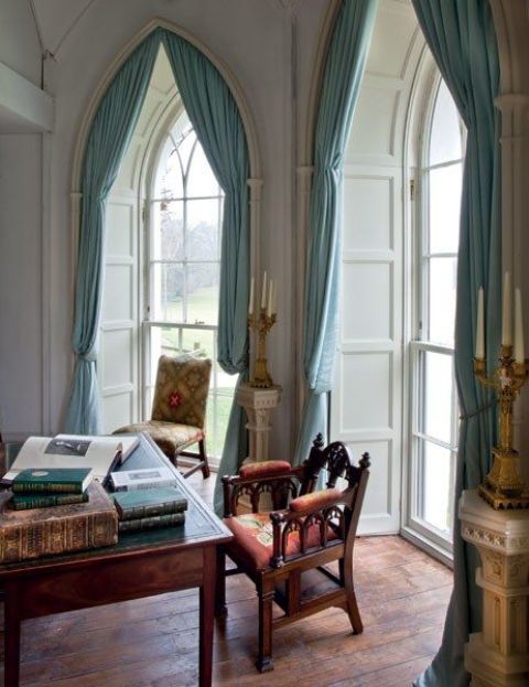 a light-colored home office with arched windows, blue curtains, a dark stained desk and heavy chairs plus candles