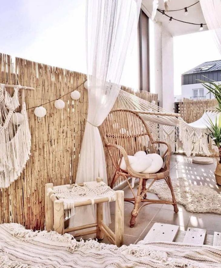 a lovely neutral boho balcony with rattan furniture, a hammock, a loveseat with a boho blanket, paper lamps, macrame and fringe is a very cozy space