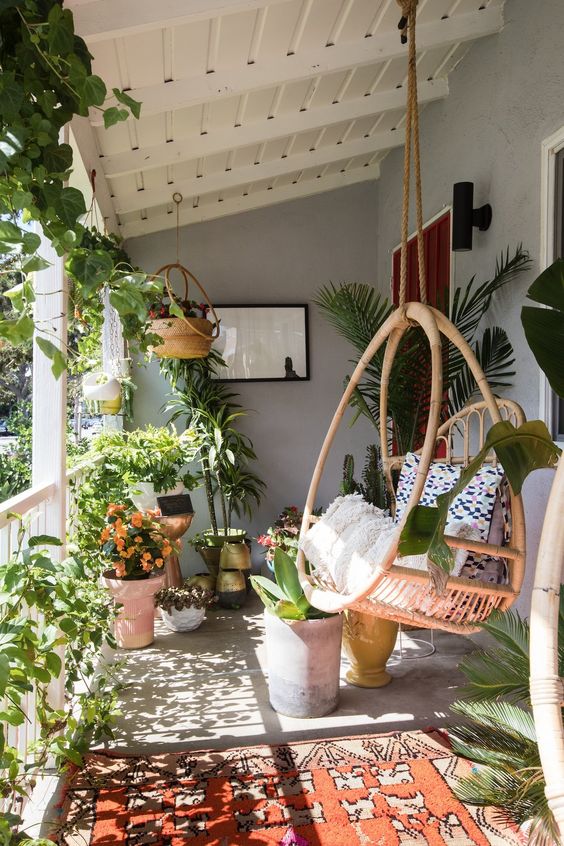 a lush boho porch with lots of greenery and blooms planted, a rattan hanging chair, a pritned rug is very cool