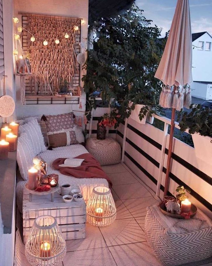 a neutral boho balcony with a daybed with boho pillows, a white crate, lots of candles and candle lanterns, lights and some boho poufs