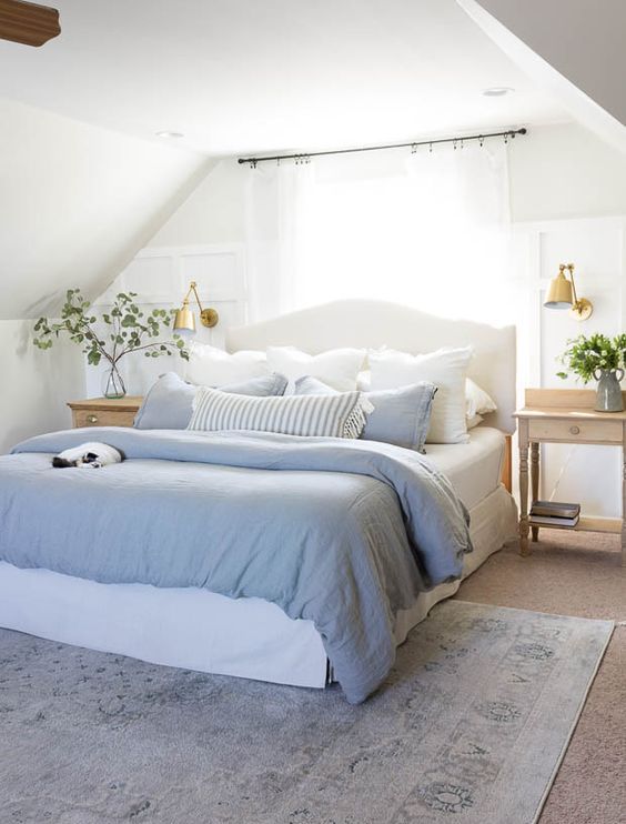 a neutral farmhouse spring bedroom with a white bed, wooden nightstands, blue and white bedding, gold sconces and greenery