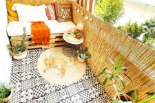 a pretty boho balcony with black and white tiles on the floor, a pallet seat with pillows, potted greenery, a jute rug and some baskets and lamps