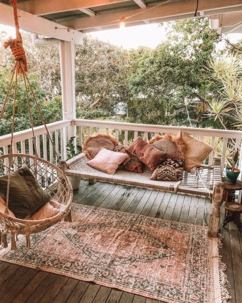 a relaxed boho porch with a woven daybed, a rattan and macrame hanging chair and a boho rug