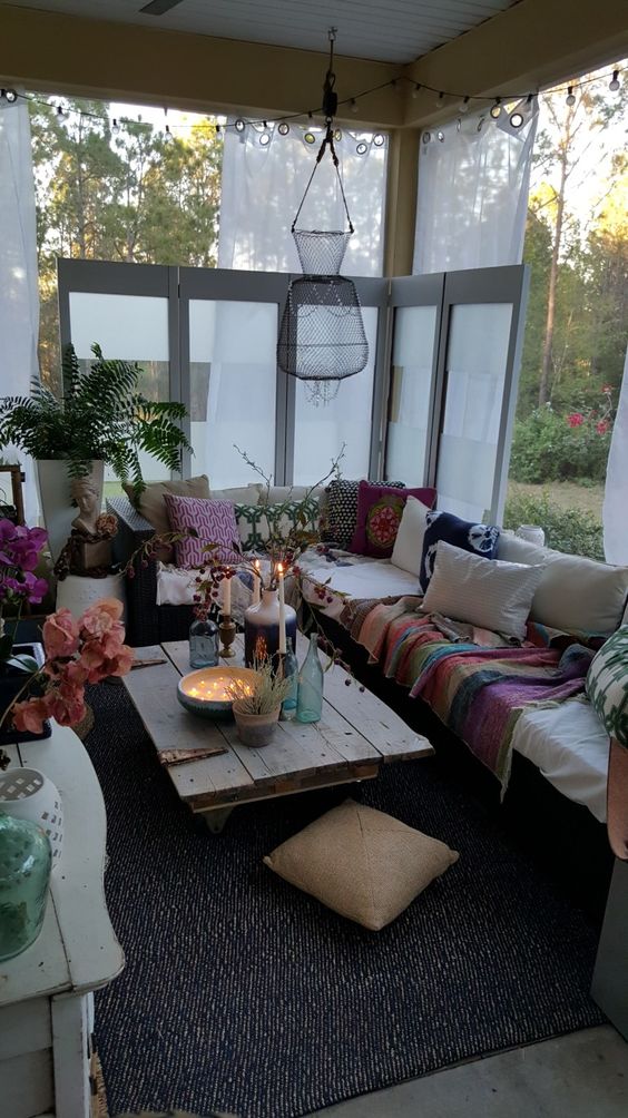 a screened boho porch with a sectional styled with bright printed textiles, a low wooden table with candles and lights and blooms