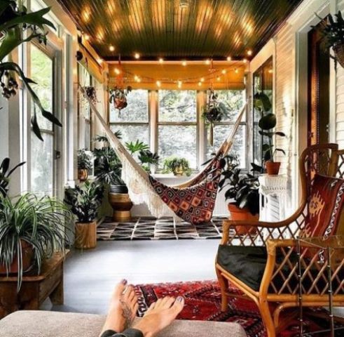 a screened porch with a macrame hammock, potted plants, lights and rattan furniture plus boho rugs