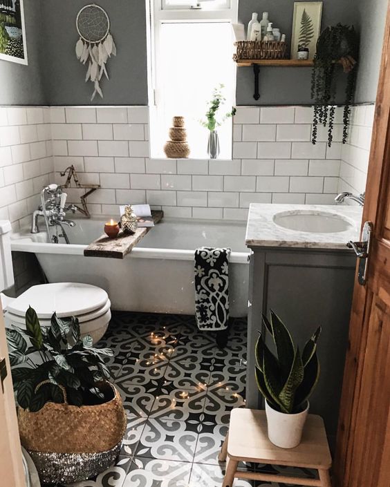 a small boho bathroom with a mosaic tile floor, grey and white tile walls, a tub, potted greenery and baskets for storage