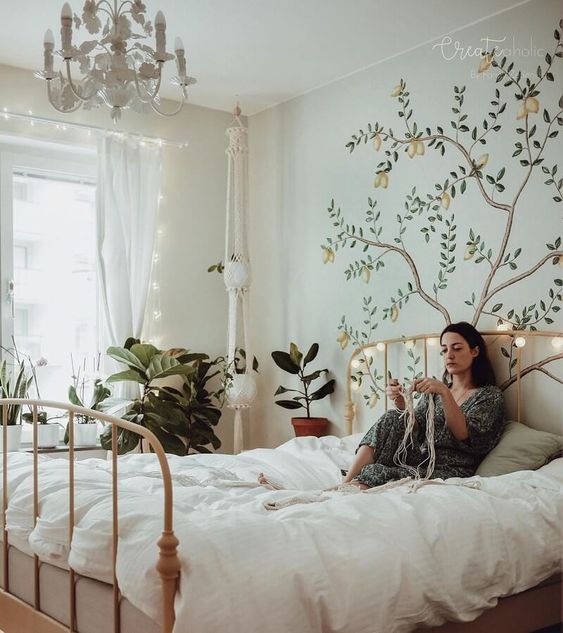 a spring boho bedroom with neutral walls, a neutral metal bed, a white chandelier, a painted tree, potted greenery