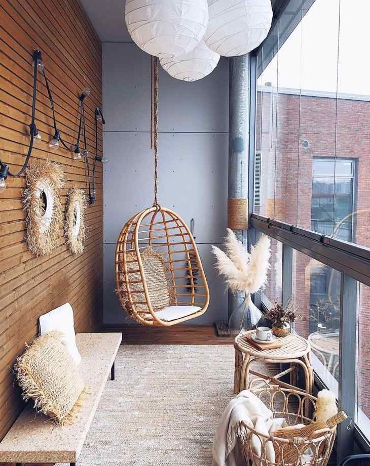 a stylish and laconic boho balcony with a jute rug, a cork bench, a rattan hanging chair, some pampas grass, rattan stools and a basket and paper pendant lamps