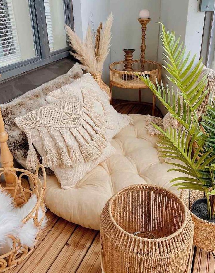 a tiny boho tropical balcony with a low seat with pillows, rattan baskets and a side table, candle lanterns, potted plants and pampas grass in a vase