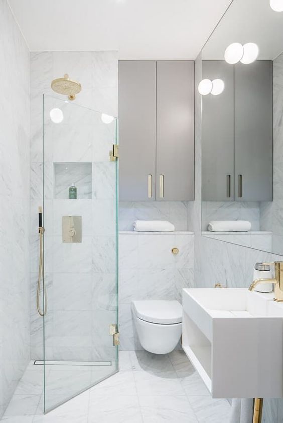 a tiny minimalist bathroom with white marble tiles, sleek grey cabinets, gilded touches and a triangle shower space